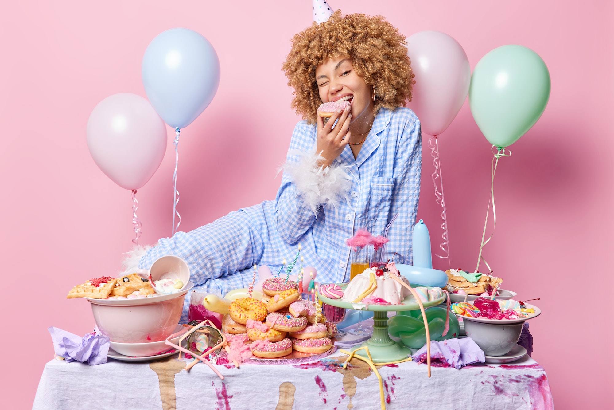 Hungry positive woman dressed in pajama bites delicious doughnut poses near messy festive table
